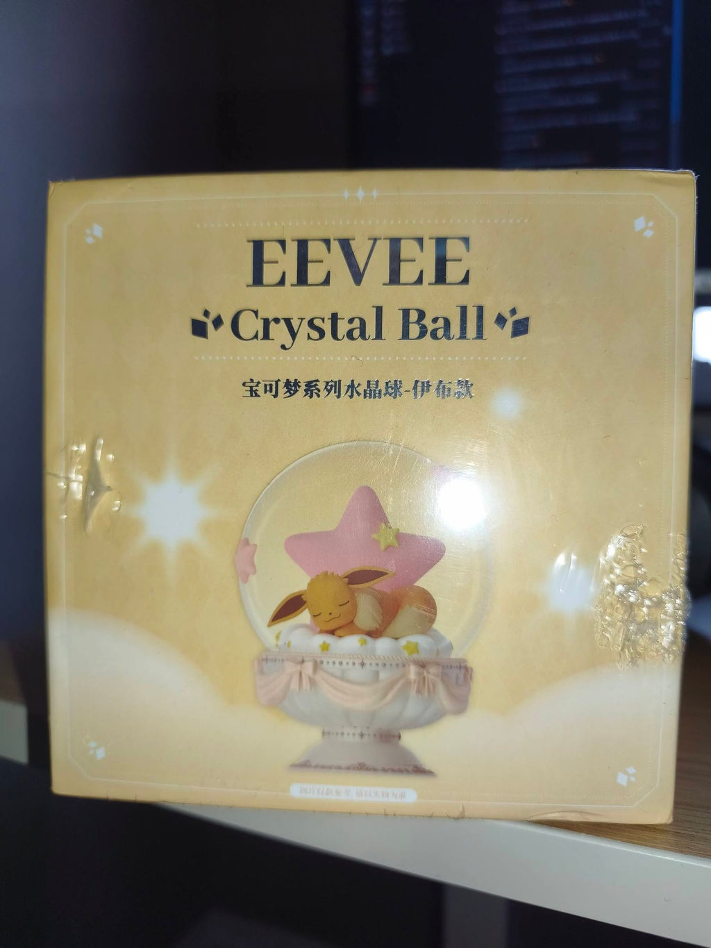 A boxed EEVEE Crystal Ball product photographed indoors on a shelf. The package is predominantly warm beige/gold with printed text reading 'EEVEE Crystal Ball' and an illustration of a pink star and a small bunny inside a clear crystal sphere.