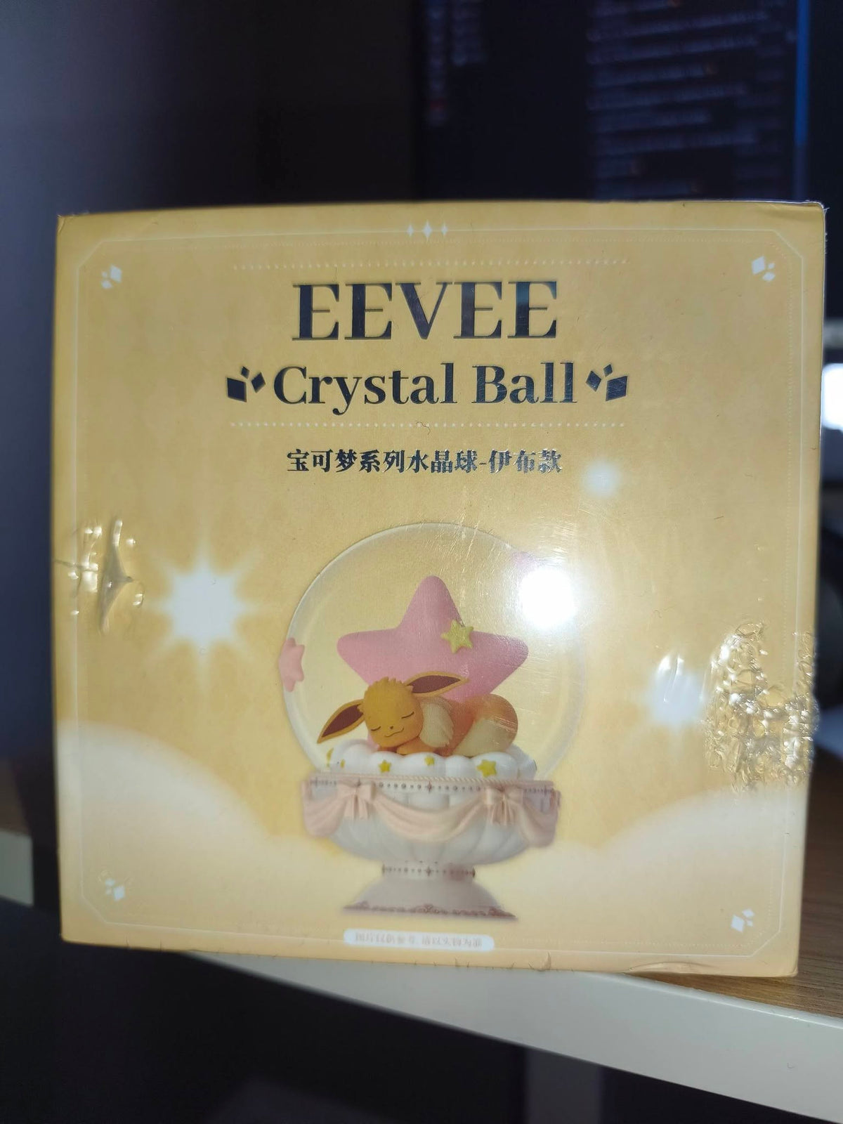 A boxed EEVEE Crystal Ball product photographed indoors on a shelf. The package is predominantly warm beige/gold with printed text reading 'EEVEE Crystal Ball' and an illustration of a pink star and a small bunny inside a clear crystal sphere.