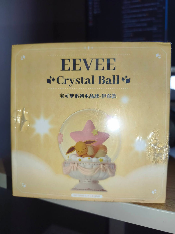 A boxed EEVEE Crystal Ball product photographed indoors on a shelf. The package is predominantly warm beige/gold with printed text reading 'EEVEE Crystal Ball' and an illustration of a pink star and a small bunny inside a clear crystal sphere.